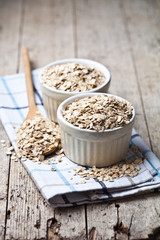 Oat flakes in ceramic bowls and wooden spoon, golden wheat ears on rustic wooden background.