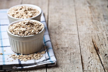 Oat flakes in ceramic bowls on linen napkin, golden wheat ears on rustic wooden background.