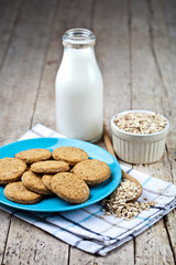 Fresh baked oat cookies on blue ceramic plate on linen napkin, bottle of milk and oak flakes on rustic wooden table.