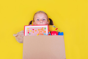 Blond little girl holding a box with toys on yellow background. Donation concept..