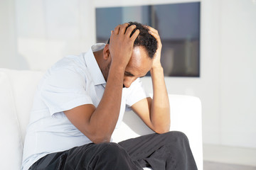 African Man suffering from headache migraine pain at home on sofa. Health problem, stress and depression. Male holds head with hand. Concept of health.