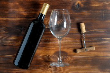 A dark bottle of wine next to a corkscrew and a twisted cork, glass goblet on a wooden background.