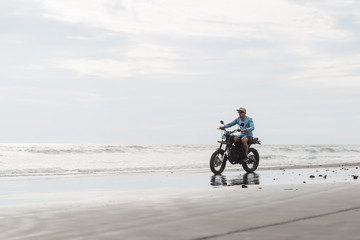 Obraz premium Man in cap riding motorcycle on beach. Moto cross dirtbiker on beach sunset on Bali. Young hipster male enjoying freedom and active lifestyle, having fun on a bikers tour.