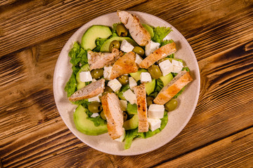Fresh salad with chicken meat, feta cheese, avocado, green olives and lettuce leaves in ceramic plate on wooden table. Top view