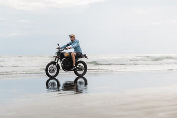 Obraz na płótnie Canvas Man in cap riding motorcycle on beach. Moto cross dirtbiker on beach sunset on Bali. Young hipster male enjoying freedom and active lifestyle, having fun on a bikers tour.