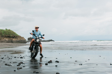 Man in cap riding motorcycle on beach. Moto cross dirtbiker on beach sunset on Bali. Young hipster...