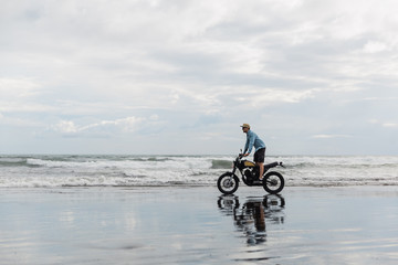 Fototapeta premium Man in cap riding motorcycle on beach. Moto cross dirtbiker on beach sunset on Bali. Young hipster male enjoying freedom and active lifestyle, having fun on a bikers tour.