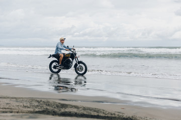 Fototapeta na wymiar Man in cap riding motorcycle on beach. Moto cross dirtbiker on beach sunset on Bali. Young hipster male enjoying freedom and active lifestyle, having fun on a bikers tour.