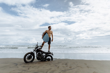 male person looking for inspiration while standing on sportive motorbike. holding surfboard in hands. Young man surfer enjoying recreation near to ocean.