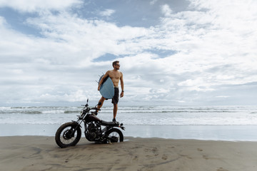 male person looking for inspiration while standing on sportive motorbike. holding surfboard in...