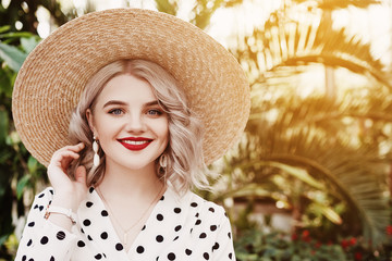 Outdoor close up fashion portrait of young beautiful happy smiling lady wearing  trendy shell earrings, stylish straw wide brim hat, polka dot blouse, posing in street with palms. Copy, empty space 