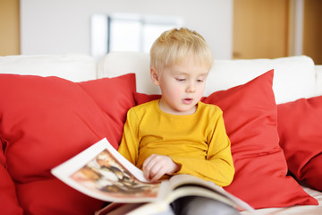 Little boy is sitting at home on the couch and reading a book. Learning to read.