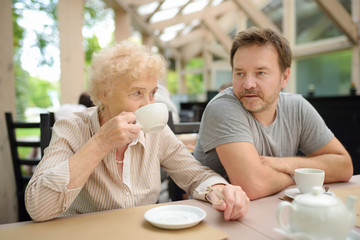 Beautiful senior lady with his mature son drinking tea in outdoors cafe or restaurant. Elderly lady lifestyle.