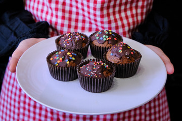 Girl holding muffins with chocolate. Woman with cakes in a candy coating. Plate with chocolate muffins on the background of a girl in an apron.