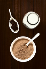 akaopulver in Schüssel mit Löffel, Schokolade und Milch an der Seite, fotografiert mit natürlichem Licht (Selektiver Fokus, Fokus auf den Kakaopulver)