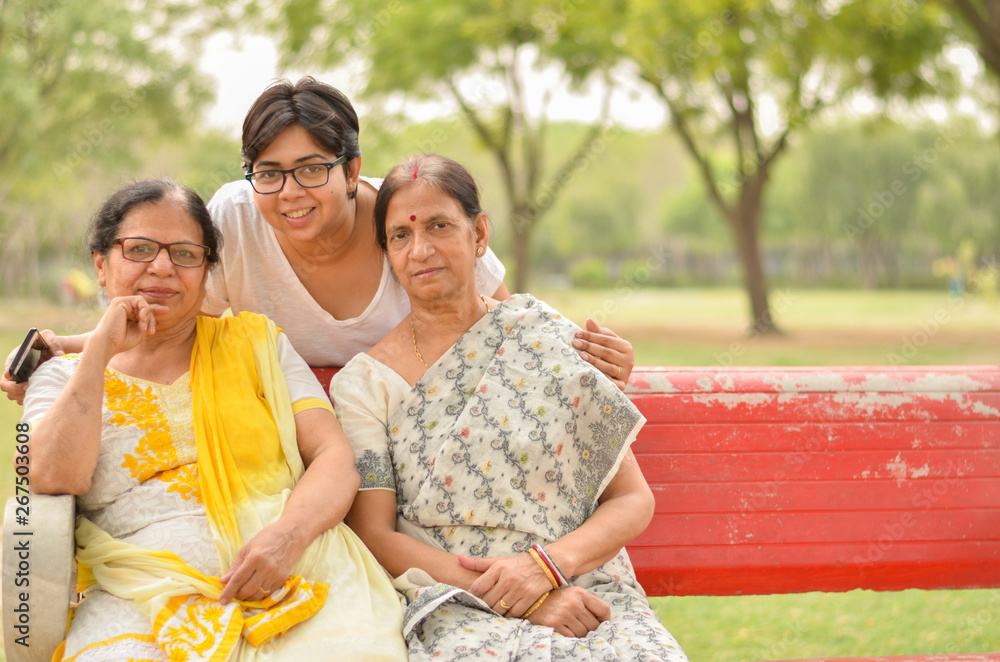 Wall mural happy looking young indian woman with two senior indian mother sitting on a red bench in a park in d