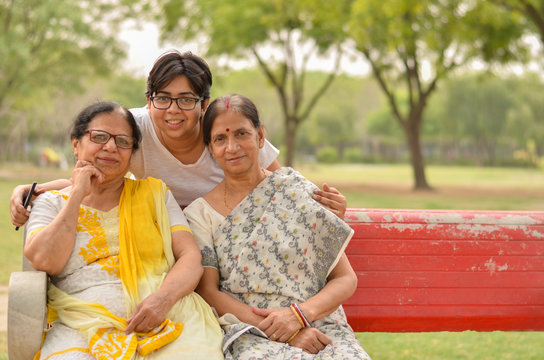 Happy looking young Indian woman with two senior Indian mother / mother in law sitting on a red bench in a park in New Delhi, India. Concept Mother's day
