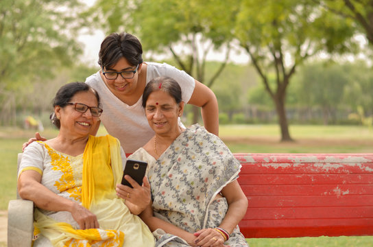 A young Indian girl with two senior women looking at the mobile phone screen while sitting on a red park bench in New Delhi, India. Concept Mother's day