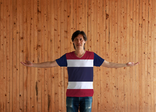 Man wearing Thailand flag color shirt and standing with arms wide open on the wooden wall background.