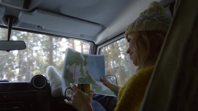 Rare view of young Caucasian woman sitting in car with map in her hands and drinking tea from steel cup
