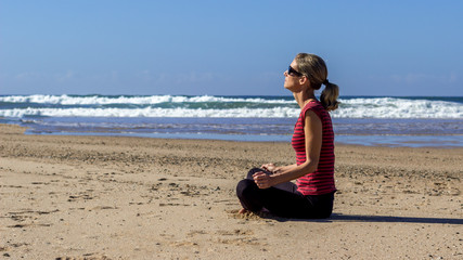 Fototapeta na wymiar young healthy woman sitting on the beach meditating in the sun with the ocean in the background