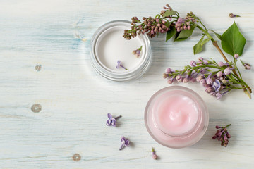 Obraz na płótnie Canvas Round glass jar with cream for skin care of the face and a branch of blossoming lilac on a light wooden background. Selective focus.