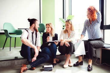 A group of young business people sitting on the floor in an office, talking.