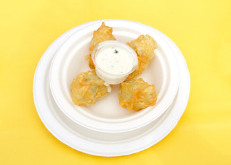 White paper plate with battered and fried artichoke with dipping sauce on a yellow table cloth. Popular street fair food.