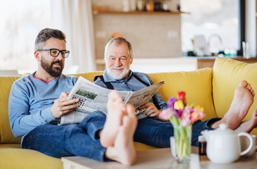 An adult hipster son and senior father sitting on sofa indoors at home, talking.