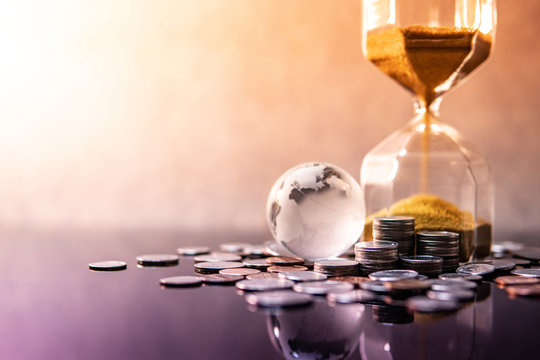 Global Business Investment Growth. Wealth Concept. Gold Sand Running Through The Shape Of Modern Hourglass With World Globe Crystal Glass And US Dollar Coins On Purple Reflective Table.