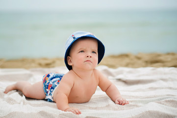 Half-year boy rest on the seafront. He lie on the bedspread on the sandy beach. Behind the back, see the surfboard. He are dressed in charming hat and bright shorts.