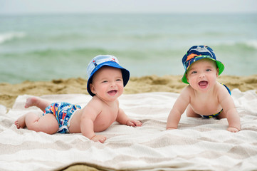 Half-year twins rest on the seafront. They lie on the bedspread on the sandy beach. Behind the back, see the surfboard. They are dressed in charming hats and bright shorts. - Powered by Adobe