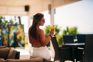 Beautiful young woman wearing swimsuit drinking a colorful cocktail sitting on a cabin of the beach club bar. Stunning girl enjoying her drink on summer time