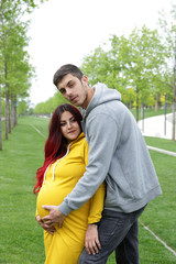 cute young man wearing a hoodie hugs his pregnant woman wearing a hoodie from behind gently with love on a green lawn in the park. Affectionate couple