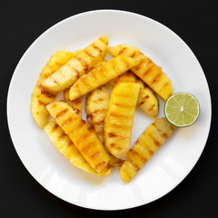 Grilled pineapple wedges with lime on a white plate over black surface, close-up. Summer food. Top view, overhead, from above.