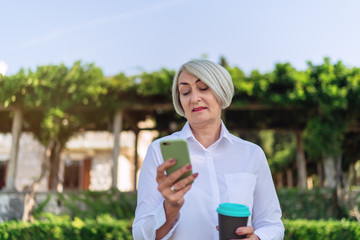 Mature woman using on mobile phone while resting at the park. Senior lady holding takeaway coffee