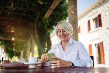 Happy mature woman using mobile phone while sitting at a cafe and drinking coffee 