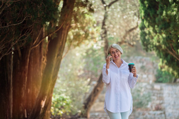 Mature woman talking on mobile phone while resting at the park. Senior lady holding takeaway coffee