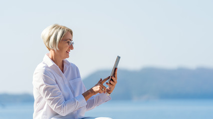 Mature woman in eyeglasses using tablet on background the sea and blue sky