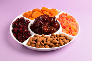 Mix of dried fruits and nuts on a white plate. Apricot, almond, raisin, dates fruit. On a lilac background. Space for text or design.