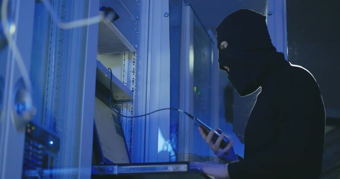 Faceless unrecognizable person in black mask using mobile phone to steal data in server room