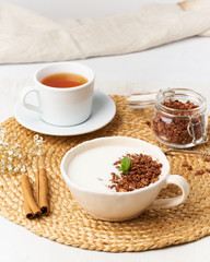 Yogurt with chocolate granola in cup, breakfast with tea on beige background, side view, vertical.
