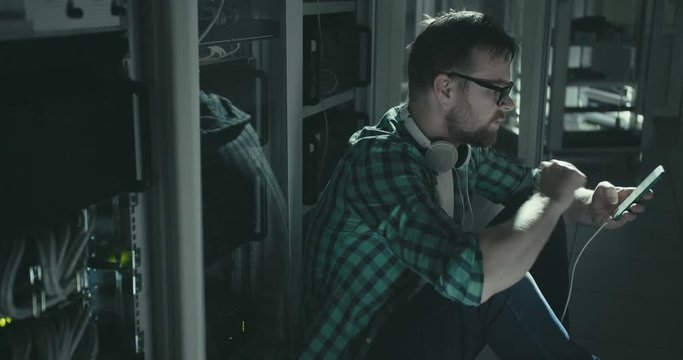 Network administrator in glasses with smartphone and earphones sitting and working in rack cabinet