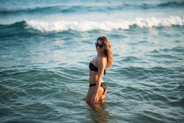 Fototapeta na wymiar Beautiful lady is waist-deep in the sea. Sexy woman in black bikini wait for waves in the sea