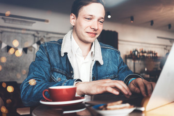Fototapeta premium Young smiling man typing message on laptop keyboard. Sitting in city cafe in smart casual dress