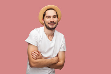 Portrait of happy successful bearded young man in white shirt with hat standing, crossed arms and looking at camera with toothy smile smile. indoor studio shot, isolated on pink background copyspace.