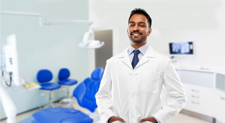 medicine, dentistry and profession concept - smiling indian male dentist in white coat over dental clinic office background