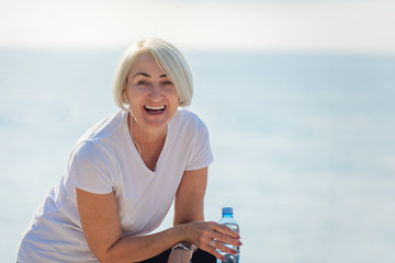 Portrait of a mature smiling woman after jogging. Senior lady drinking water  on background the sea...