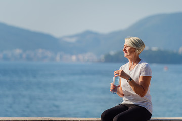 Mature woman drinking water after jogging on background the sea and blue sky