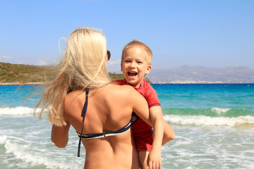 Naklejka premium Young slim woman holding her son on the beach by the clear blue sea. Little boy in UV filter clothes is smiling, mother and her cute child are having fun, holiday with children concept